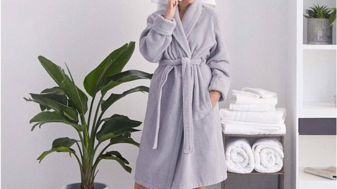 A person wearing a plush light gray bathrobe with a waist tie stands in a bright, modern bathroom beside neatly folded white towels and a large green indoor plant, creating a clean and spa-like atmosphere.