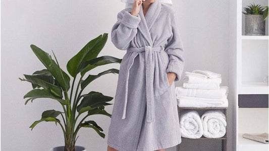 A person wearing a plush light gray bathrobe with a waist tie stands in a bright, modern bathroom beside neatly folded white towels and a large green indoor plant, creating a clean and spa-like atmosphere.