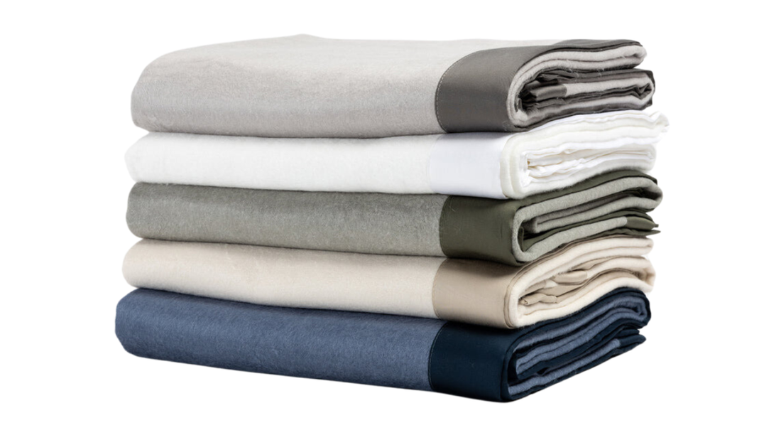 A neatly stacked collection of folded bed sheets in assorted neutral colors, including gray, white, beige, olive, and navy, displayed against a clean white background to highlight their soft texture and minimalist design.